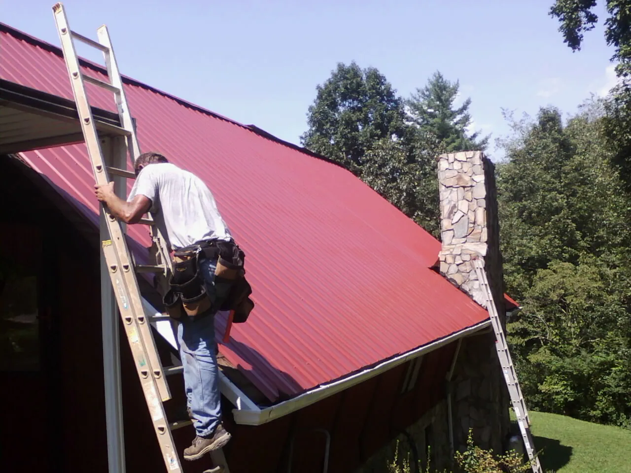 Roofer on a ladder beside a red metal roof and stone chimney — Soffit & Fascia Repair by Roofing Brothers in South Whitley